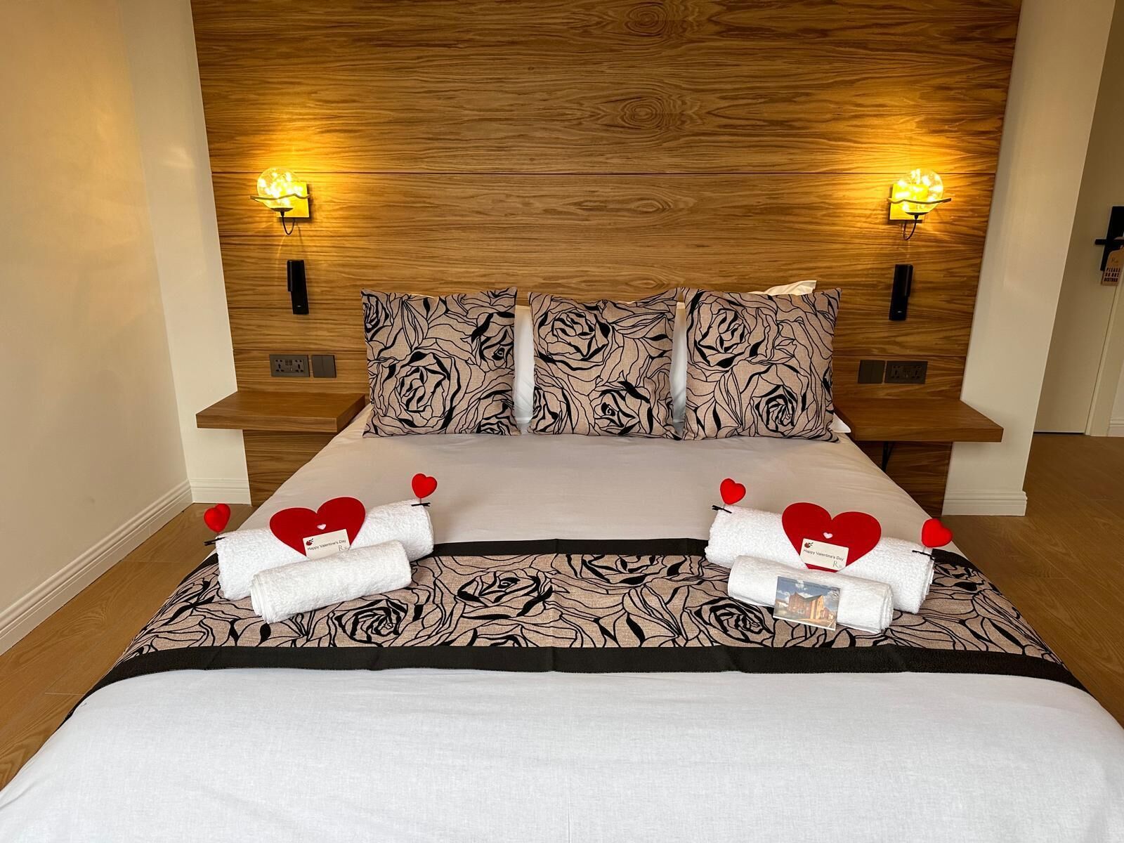 A hotel room with a wooden headboard, two bedside lamps, floral-patterned pillows, a bed with a white and black bedspread, and two towels with red heart decorations.