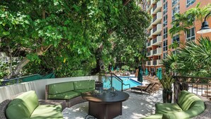 A rooftop patio with wicker furniture, a central fire pit, and a pool in the background.