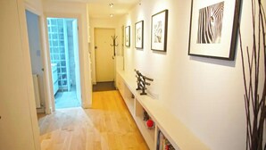 A hallway with a wooden floor, a white wall with framed artwork, and a bookshelf with books. - Superior Canary Wharf  O2 Arena 2Bed (London)