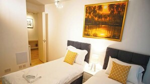 A hotel room with two single beds, a nightstand with a lamp, a painting of a sunset, and a door leading to another room. - Superior Canary Wharf  O2 Arena 2Bed (London)