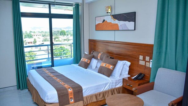 A hotel room with a large bed, a bedside table, a chair, a round table, and a view of the city through the window. - UNIQUE ROYAL HOTEL AND SUITES (Dodoma)