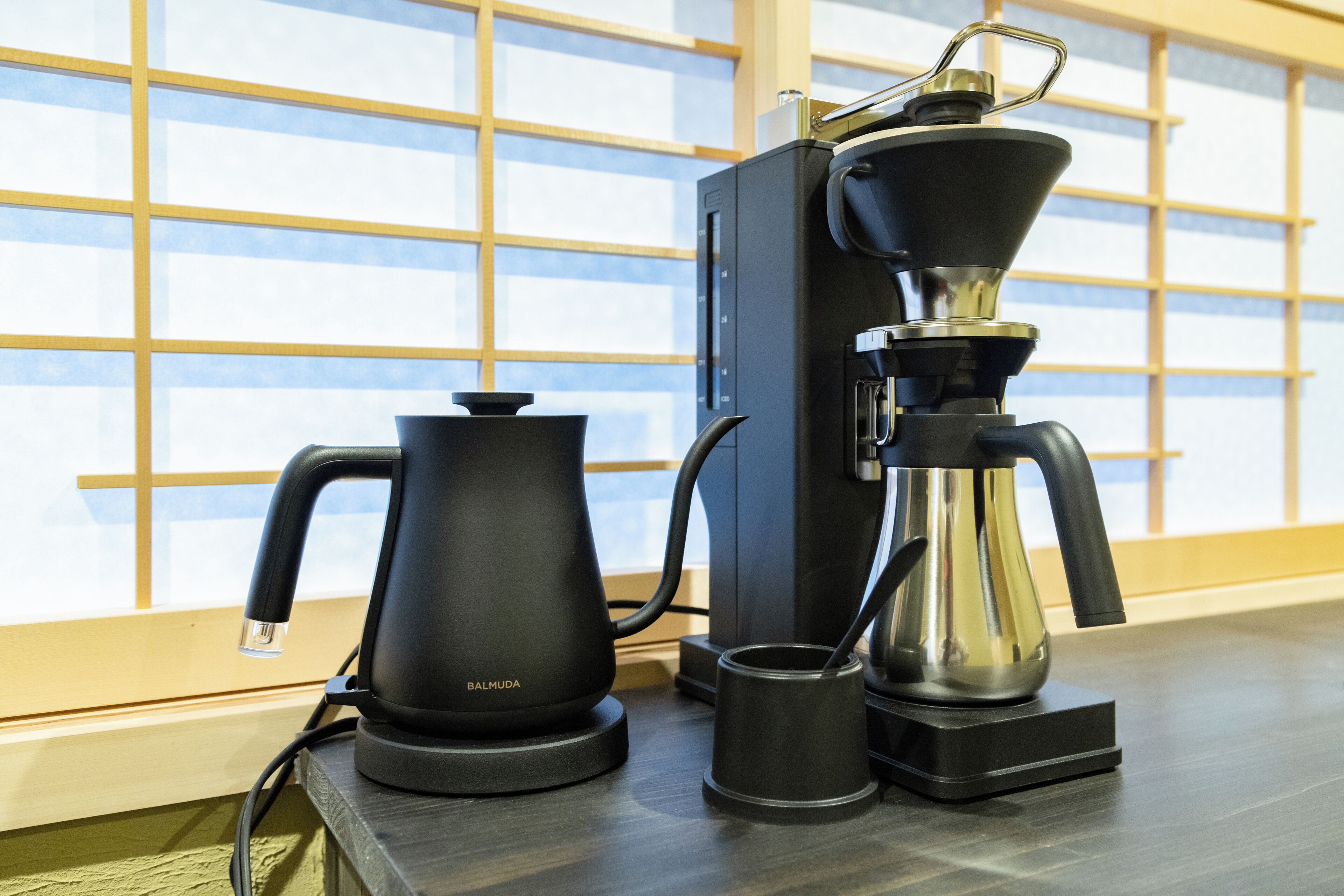 A coffee maker and kettle on a wooden table with a window in the background.