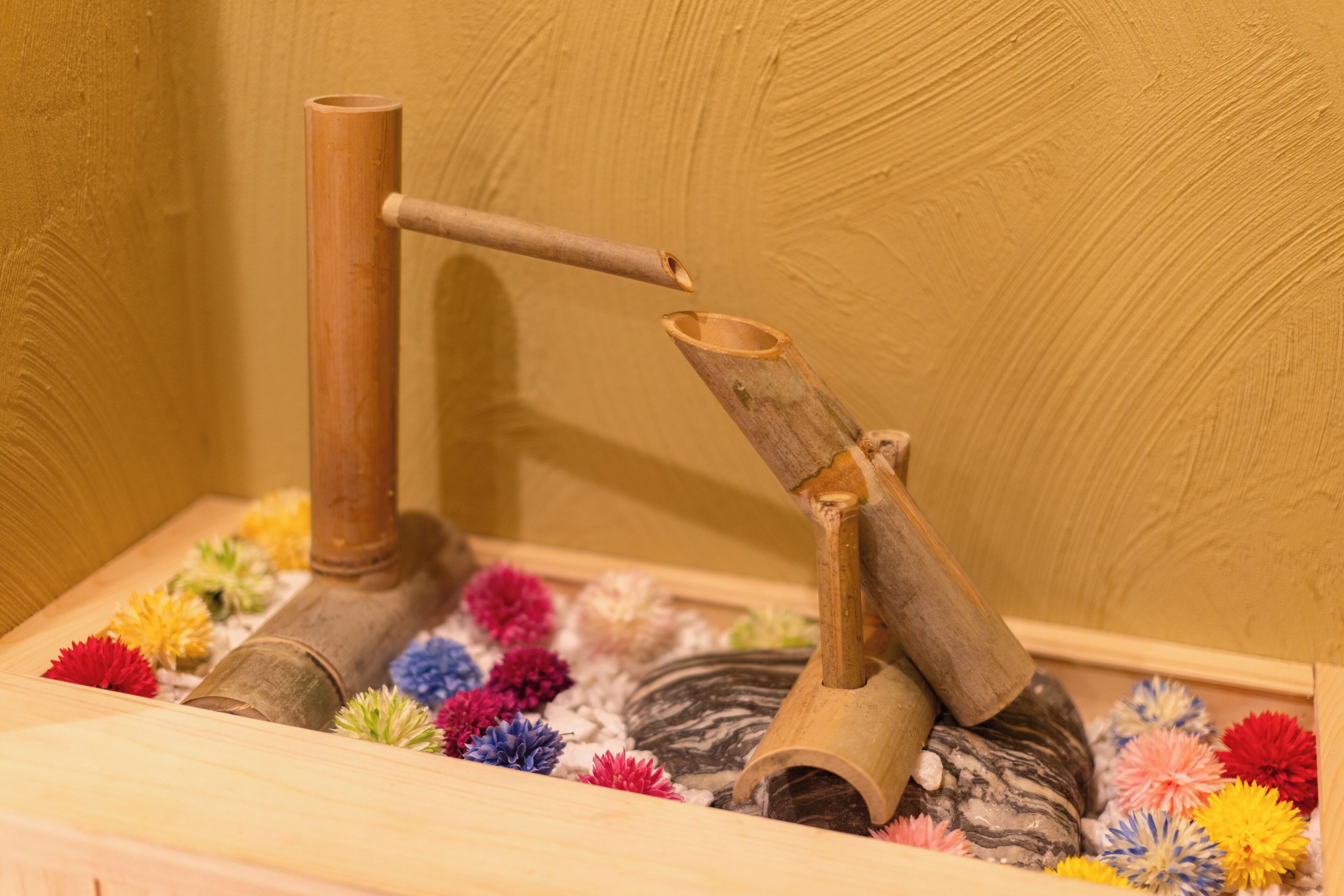 A wooden structure with a pipe and a funnel-like object, surrounded by colorful artificial flowers.