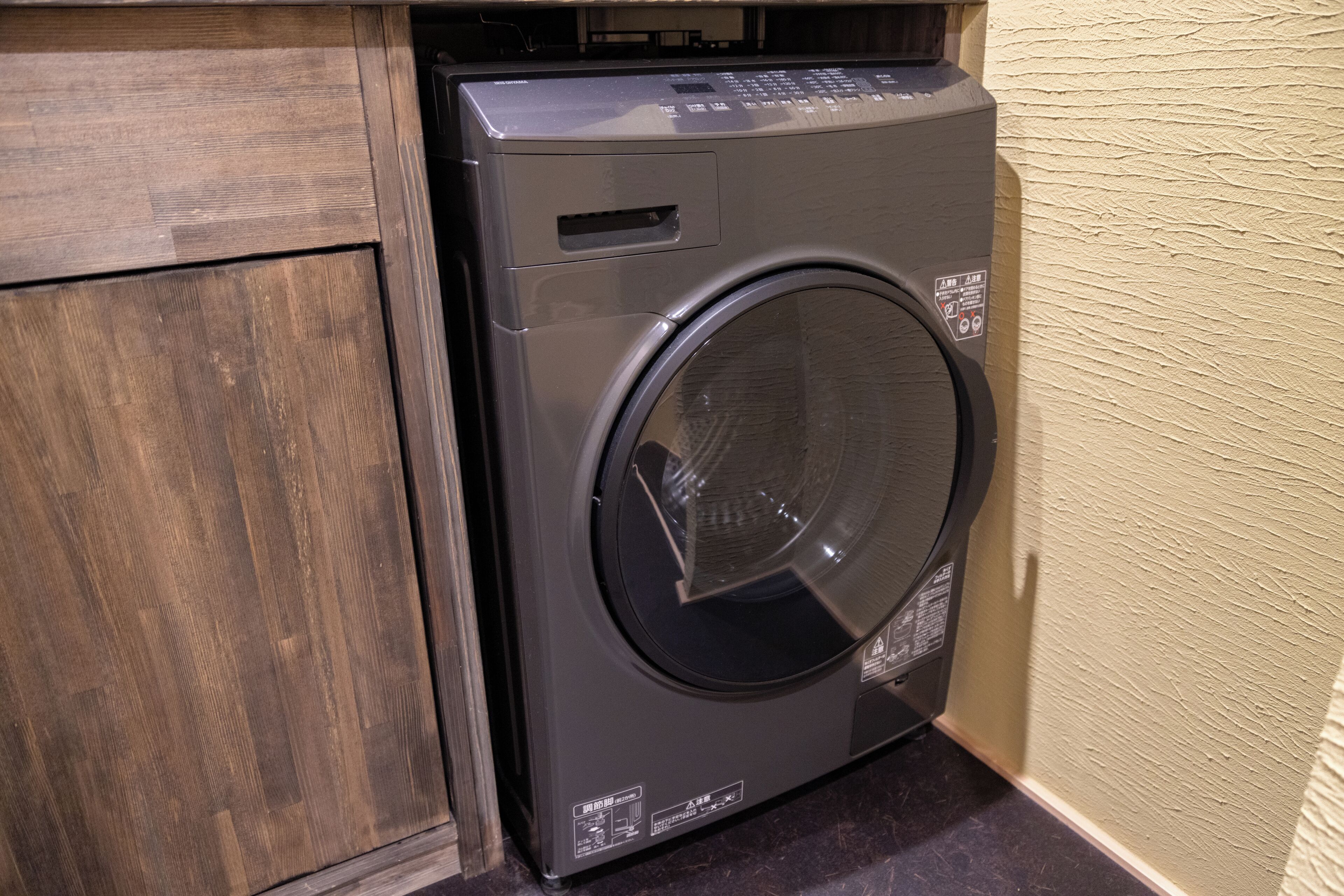 A front-loading washing machine in a corner with a wooden cabinet and a textured wall.
