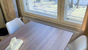 Una habitación con una mesa de madera, sillas blancas y una ventana que da a árboles y una masa de agua.