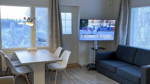 Una habitación de hotel moderna con piso de madera, una mesa de comedor con sillas, un sofá, una cama y un televisor que muestra un evento deportivo.