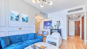 Een moderne woonkamer met een blauwe bank, een witte fauteuil, een marmeren salontafel met zakdoekjes en een schaal muffins, en een keuken op de achtergrond.