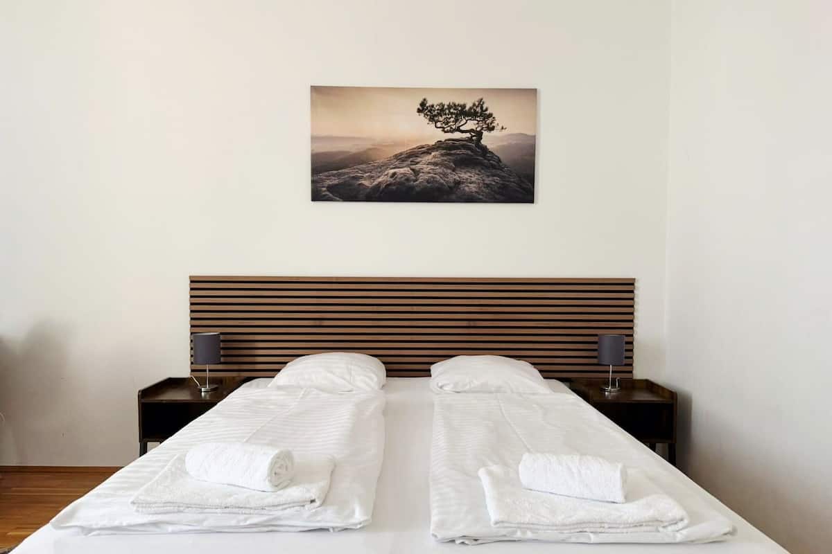 A double bed with white linens and towels, a wooden headboard, bedside tables with lamps, and a framed picture of a tree on a mountain.