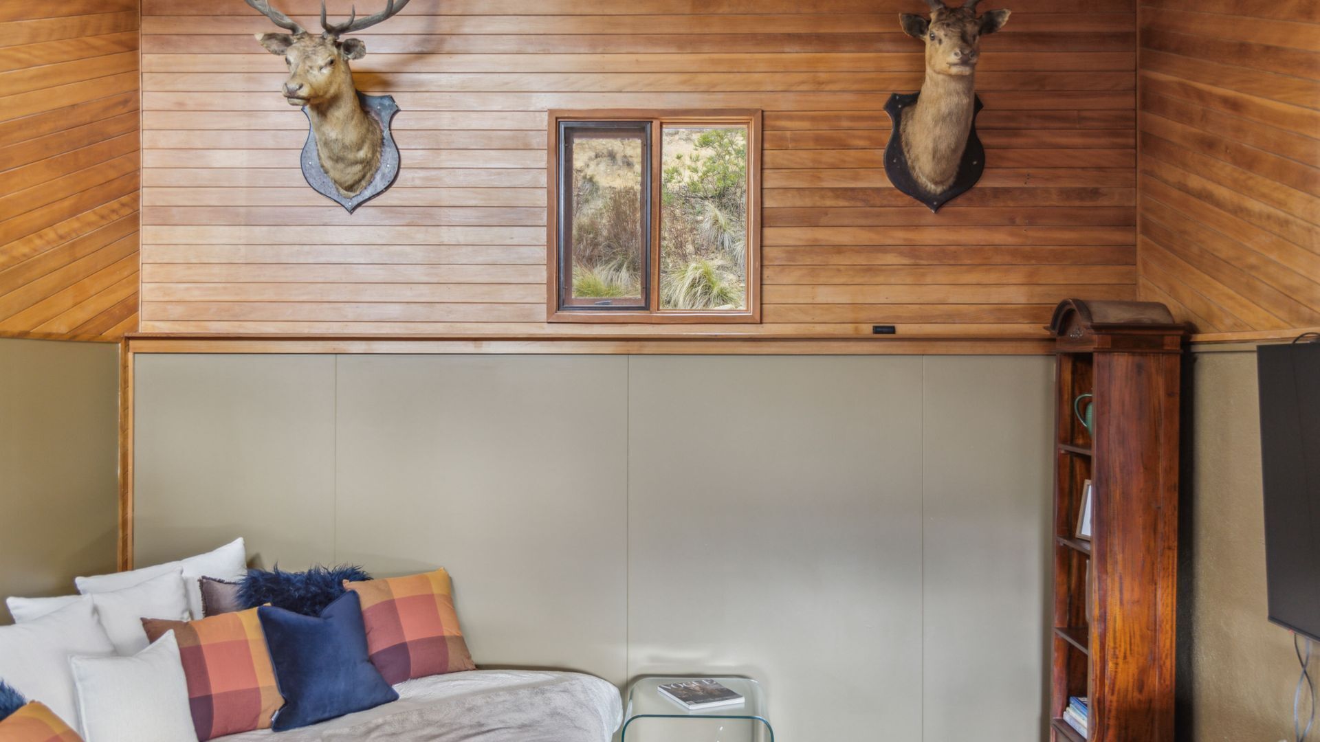 A room with wooden walls, a bed with colorful pillows, a window with a view of trees, and mounted animal heads.