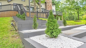 Una casa moderna con chimenea de piedra, terraza de madera y jardín ajardinado con árboles pequeños y caminos de grava.