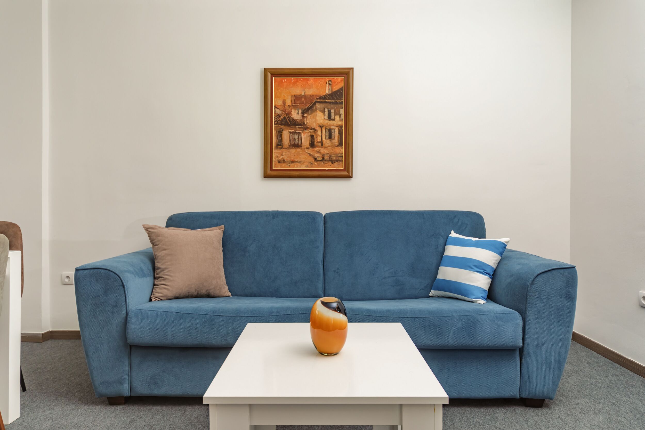 A blue sofa with two pillows, a white coffee table, and a framed painting on the wall.