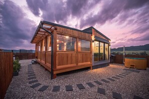Eine Holzhütte mit großen Fenstern und einem Whirlpool, umgeben von einem Kiesweg und einem Holzzaun.