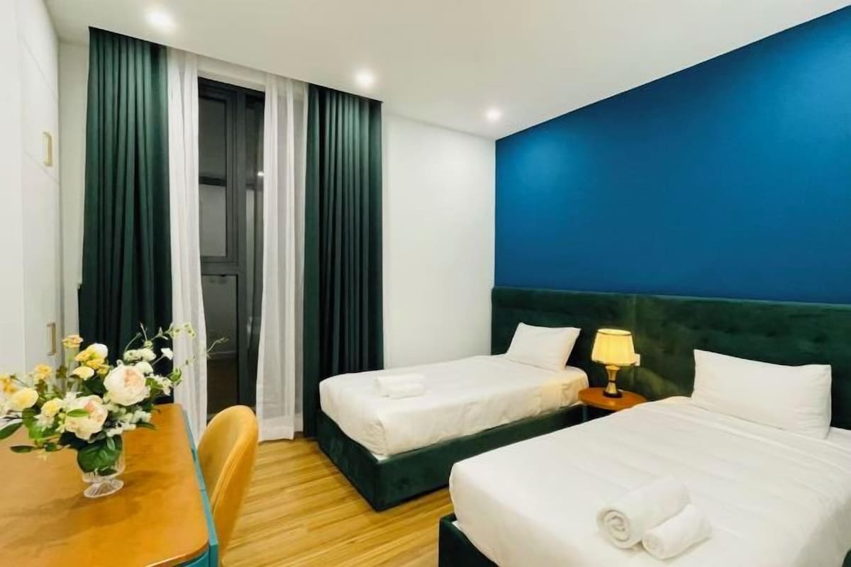 A hotel room with two beds, a wooden desk, and a vase of flowers.