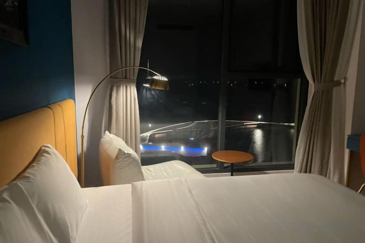 A hotel room with a bed, a nightstand, a lamp, a window with curtains, and a view of a cityscape at night.