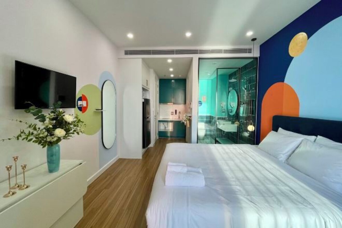 Una habitación de hotel moderna con una cama grande, un televisor de pantalla plana, un tocador con espejo y un baño visible a través de una puerta de vidrio.