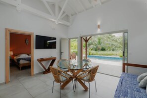 Quarto moderno com mesa de jantar de vidro, cadeiras de vime, cama, televisão e vista para uma piscina.