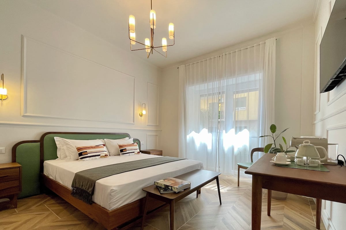 Une chambre d’hôtel avec un lit, une table de chevet, une petite table avec une théière et une fenêtre avec des rideaux.