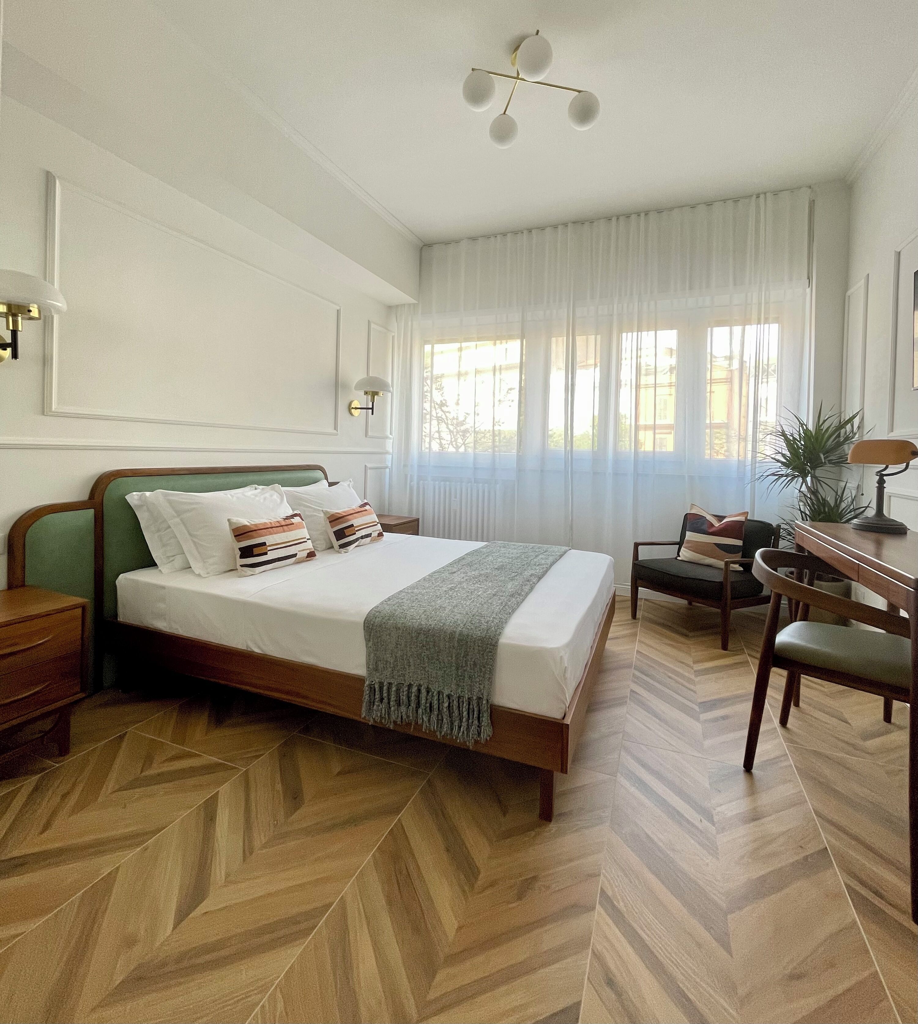 A bedroom with a wooden herringbone floor, a bed with a green headboard, a desk with a chair, and a window with sheer curtains.