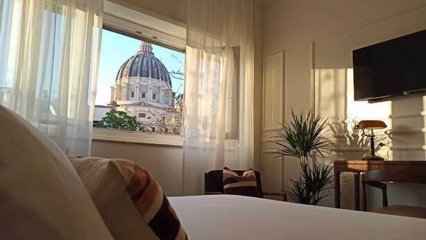A hotel room with a bed, a desk with a lamp, a TV, and a view of a large building through the window. - The New Guesthouse Vatican (Rome)