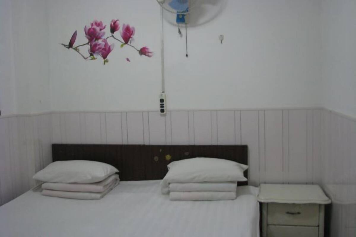 A bedroom with a bed, two pillows, a nightstand, a wall-mounted fan, and a floral decoration.