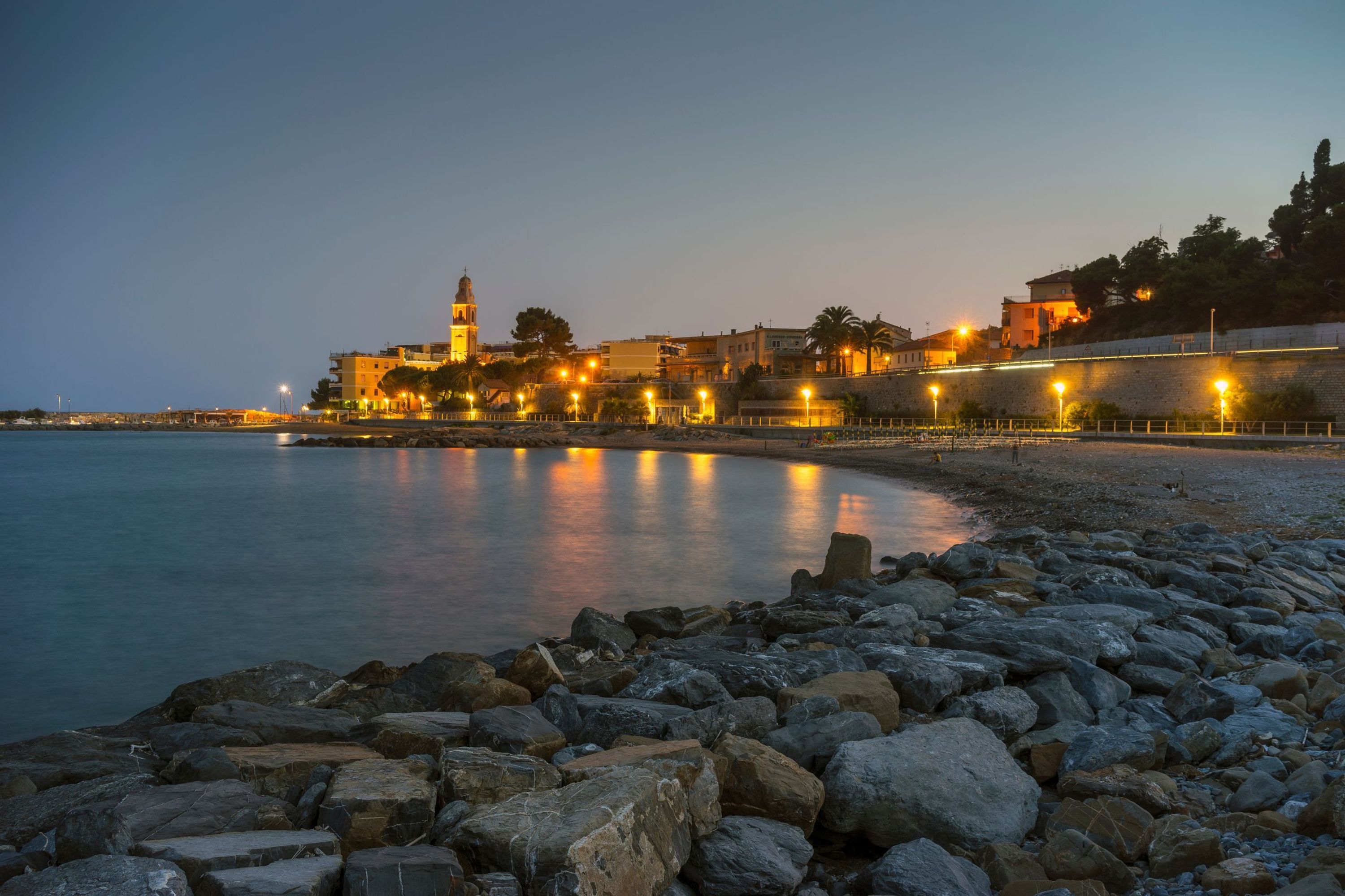 A coastal town at dusk with illuminated buildings, a prominent tower, and a rocky shoreline.