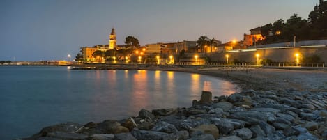 A coastal town at dusk with illuminated buildings, a prominent tower, and a rocky shoreline.
