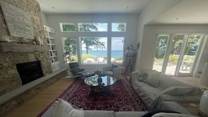 Een ruime woonkamer met een stenen openhaard, een groot raam met uitzicht op zee en een rood tapijt met een patroon.