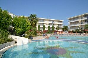 Una piscina con gente, palmeras y un edificio con balcones al fondo.