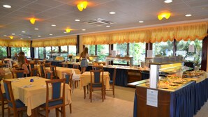 Un restaurante tipo bufé con varias mesas y sillas, una barra de bufé con diversos alimentos y ventanas grandes con cortinas.