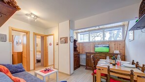 A cozy living room with a sofa and a dining table set for two, a television, and a kitchen area with wooden cabinets and a window. - La Tana Dell Ermellino 3 Ski In Ski Out (Sestriere)
