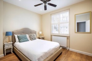 Ein Schlafzimmer mit Bett, Nachttischen, Deckenventilator, einem Fenster mit Jalousien, einem Heizkörper und einem Spiegel an der Wand.