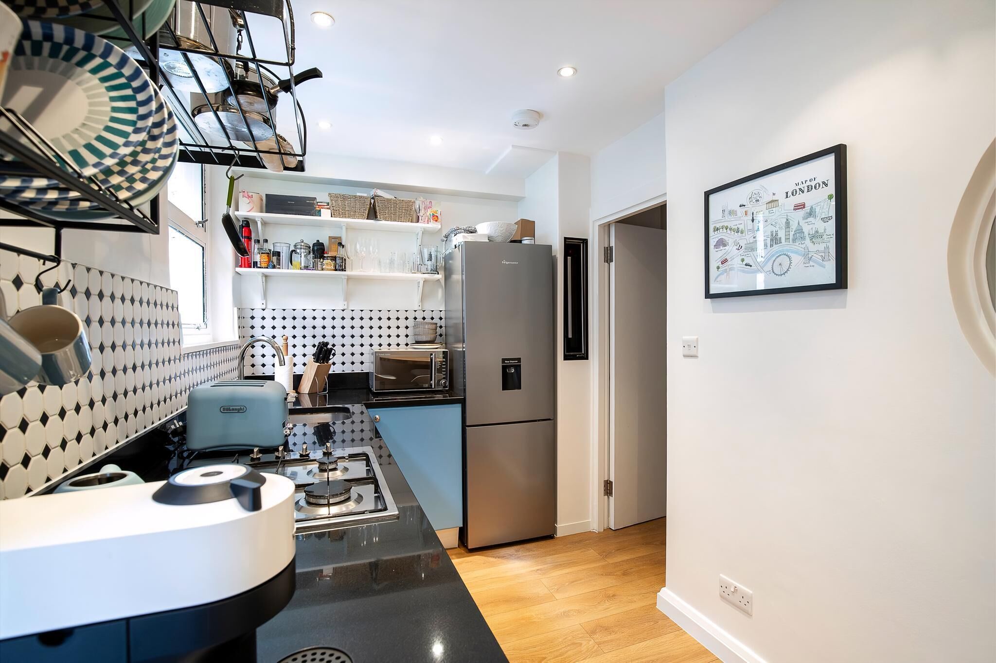 Eine moderne Küche mit Gasherd, Edelstahlgeräten und einer Mikrowelle. Ein Kühlschrank, eine Spüle und ein Regal mit diversen Gegenständen sind vorhanden. An der Wand hängt ein gerahmtes Bild von London.