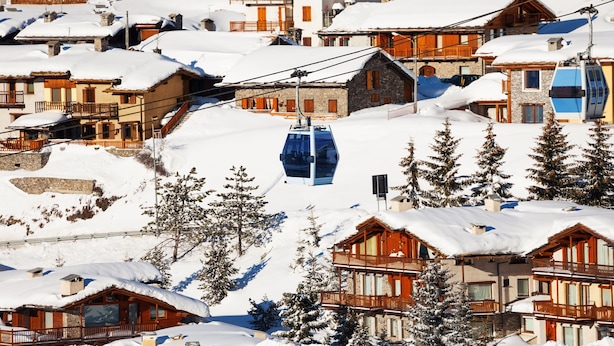 A snow-covered mountain village with wooden buildings, a ski lift, and pine trees.