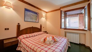 A bedroom with a wooden headboard, a bed with a checkered bedspread, two bedside tables, a wall-mounted lamp, a map, and a window with wooden shutters.