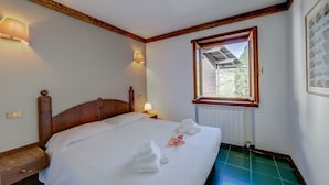 Una cama bien hecha con sábanas y toallas blancas, un cabecero de madera, una mesita de noche con una lámpara y una ventana con vistas a zonas verdes.