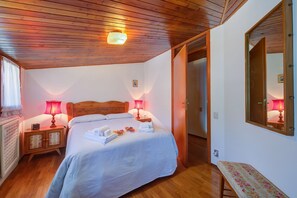 A bedroom with a wooden ceiling, a bed with white linens, two bedside lamps, a wooden nightstand, and a bench with floral upholstery. - Il Rododendro City Center (Bardonecchia)