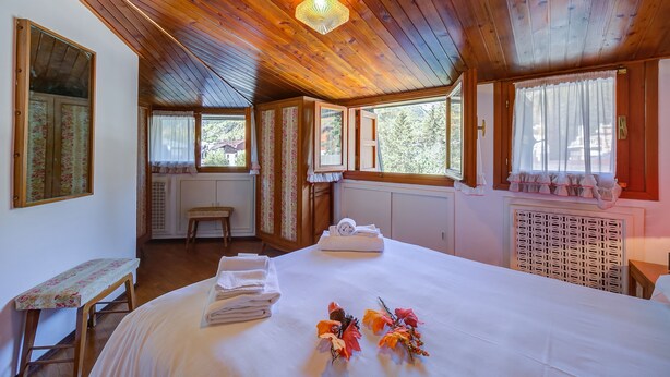 Quarto com teto de madeira, cama com lençóis brancos, um banco e janela com cortinas.
