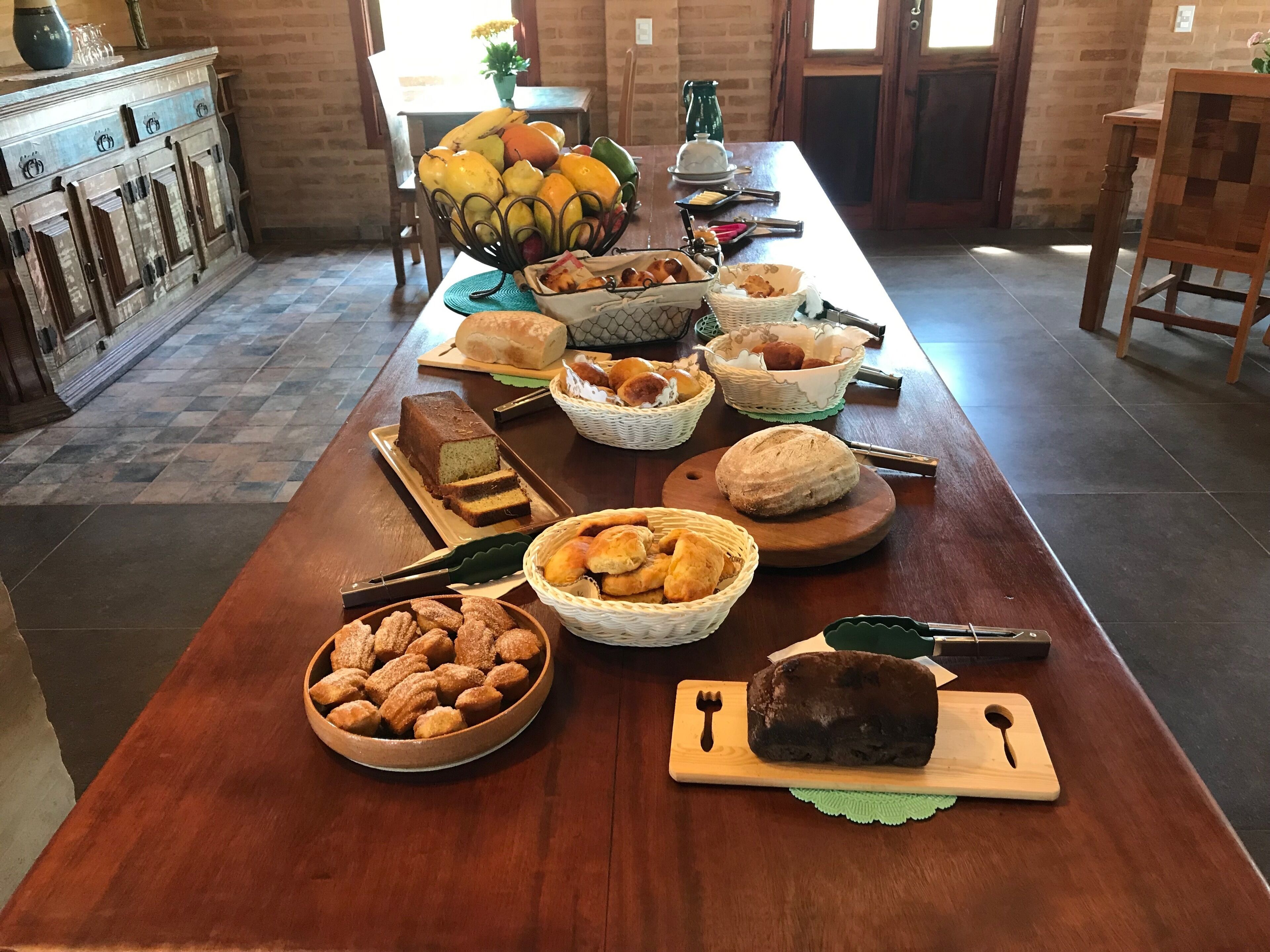 Una larga mesa de madera con diversos alimentos y utensilios, incluyendo pan, pasteles y una hogaza de pan sobre una tabla de cortar.