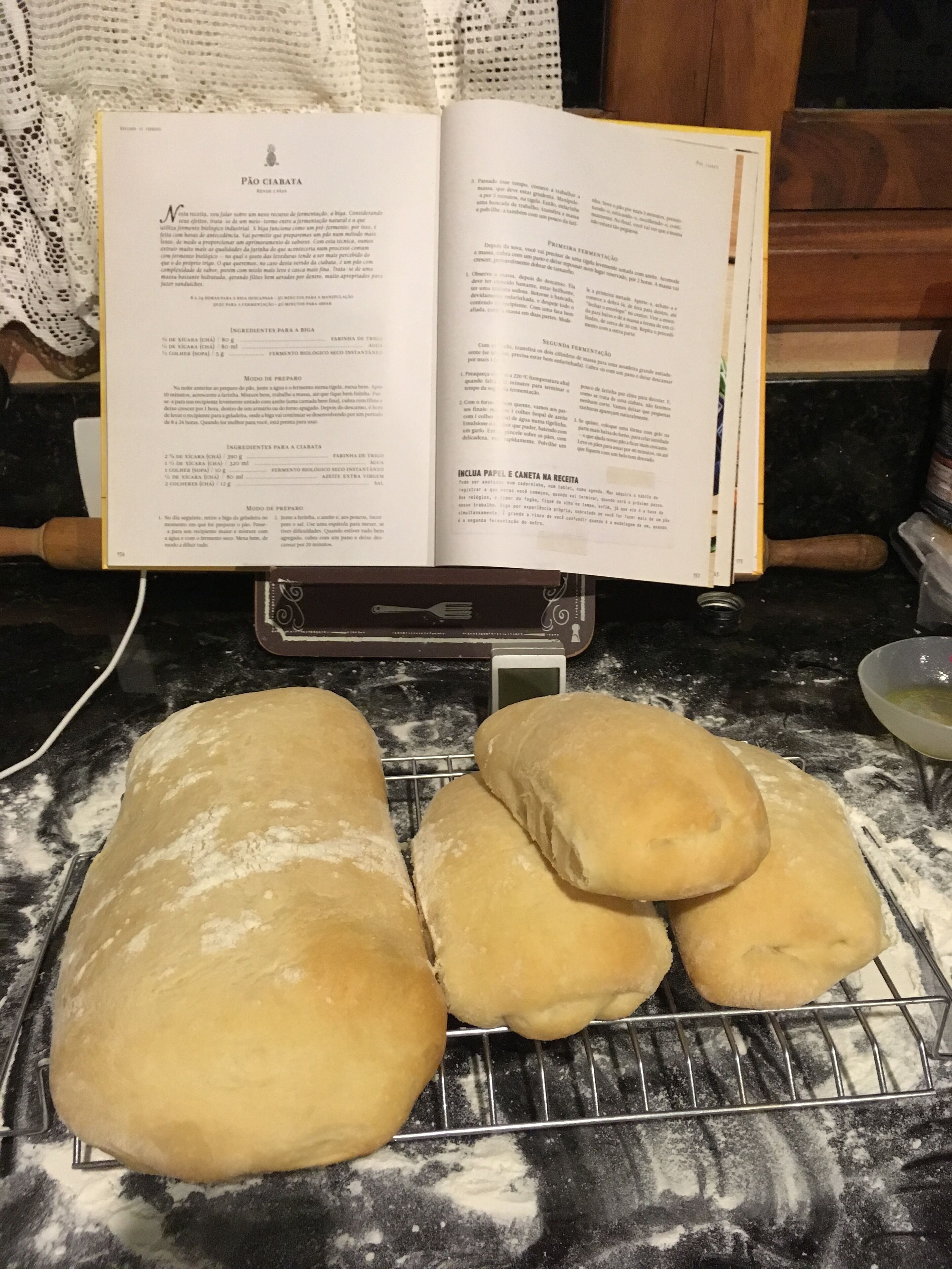 An open recipe book on a kitchen counter with dough shaped like bread rolls cooling on a rack.