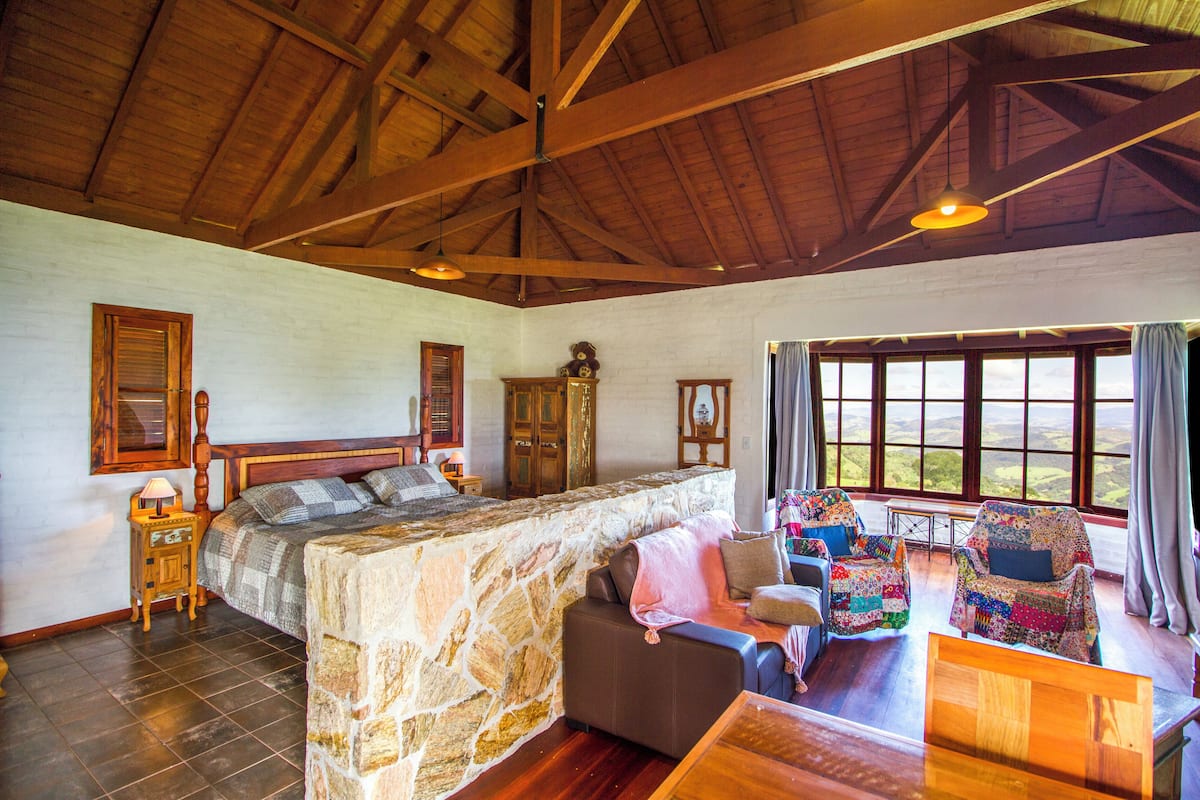 A bedroom with a stone wall, wooden ceiling beams, a bed, a dresser, and a view through large windows.