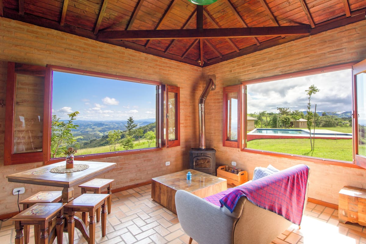 A cozy room with a fireplace, wooden furniture, and large windows offering a view of a swimming pool and lush landscape.
