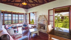 A cozy living room with a stone fireplace, large windows, and a view of the countryside. - Magnificat Cottage (Cunha)