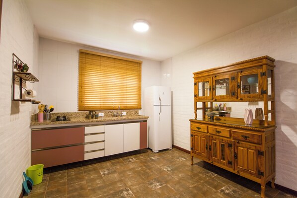 A compact kitchen with a refrigerator, wooden cabinet, and a window with blinds. - Magnificat Cottage (Cunha)