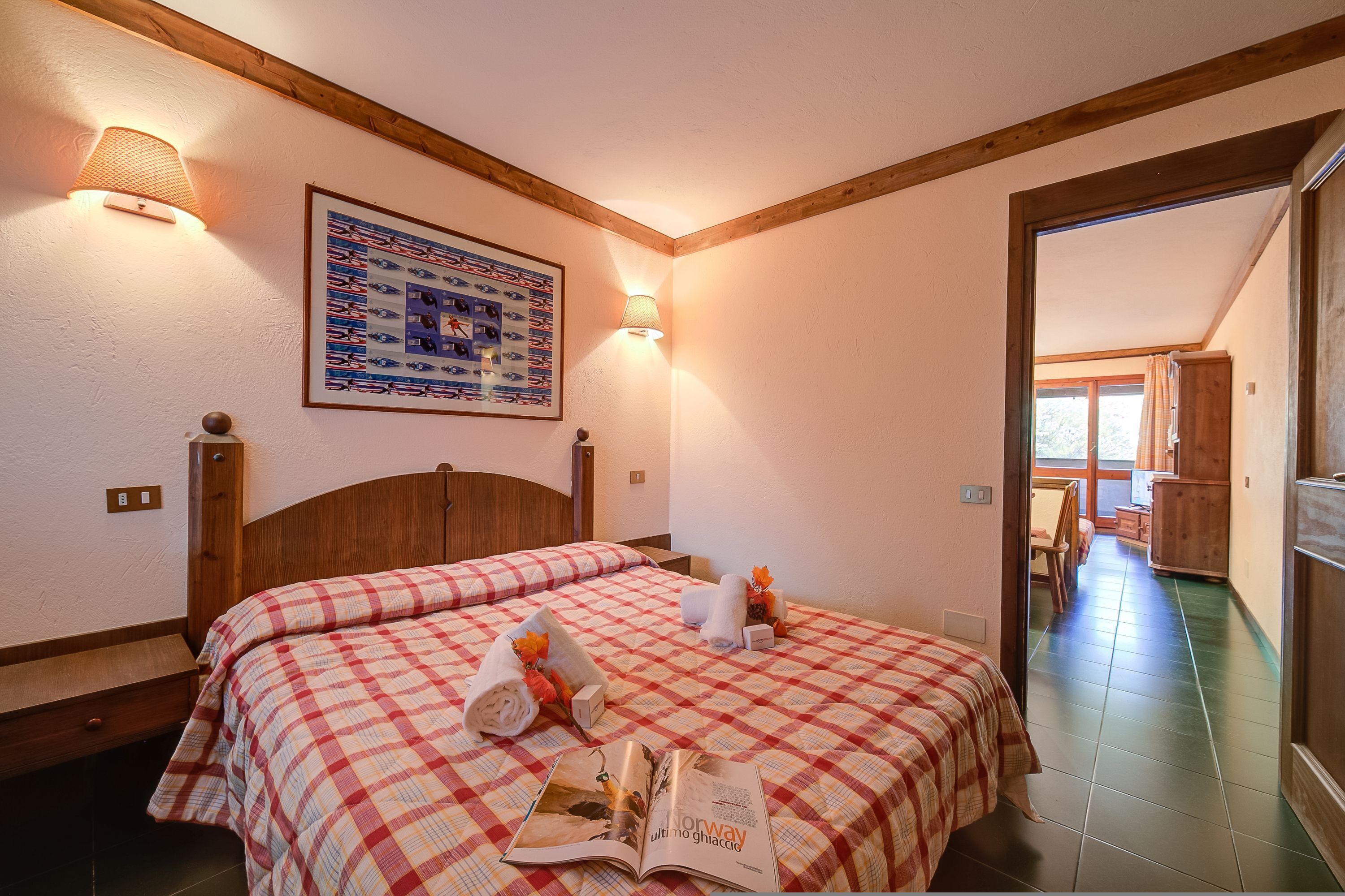 A hotel room with two beds, a wooden headboard, a wall-mounted lamp, a framed picture, and a magazine on the bed.
