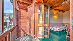 Een houten balkon met een houten hekwerk, een glazen deur die uitkomt in een kamer met een bed en een tafel, en een uitzicht op de omliggende gebouwen.