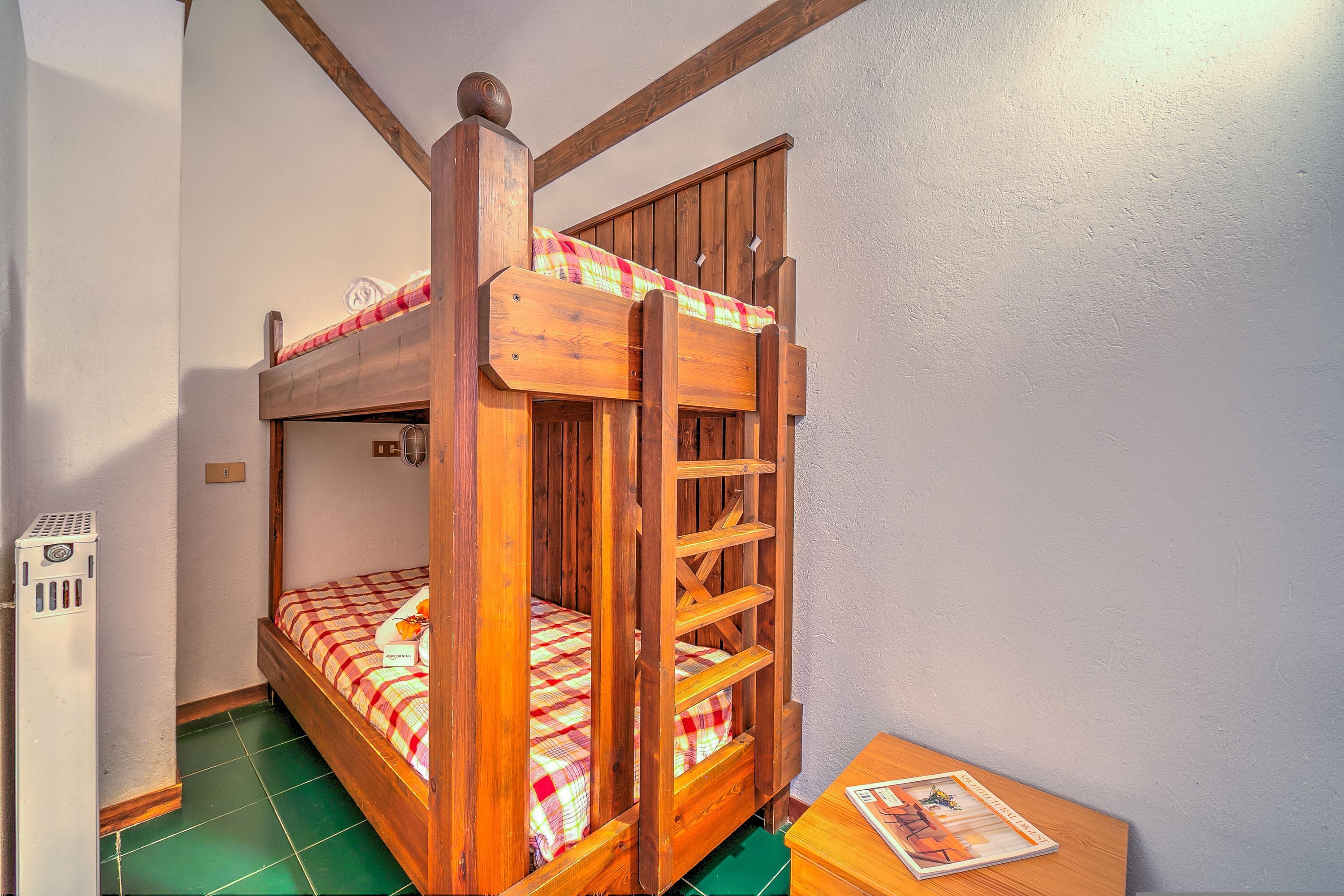Un lit superposé avec une couverture à carreaux, une échelle en bois et une petite table sur laquelle repose un magazine.