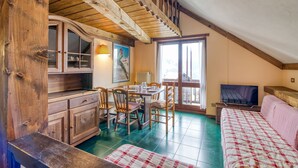 Een knus ingerichte kamer met een houten plafond, een eettafel met stoelen, een bed met een geruit plaid en een televisie.