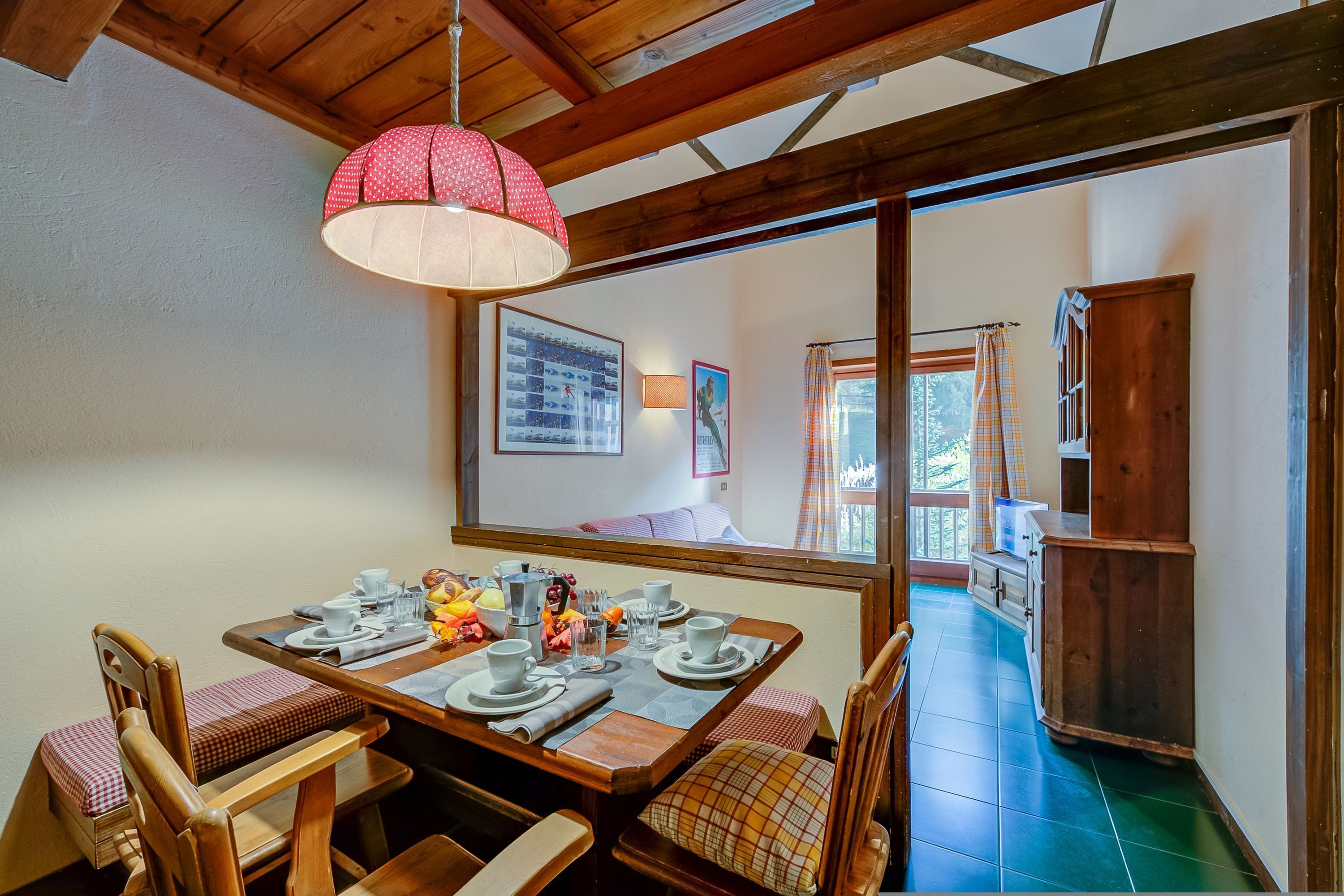 A dining area with a wooden table set for four, a red polka-dotted lamp, and a view of a balcony through a window.