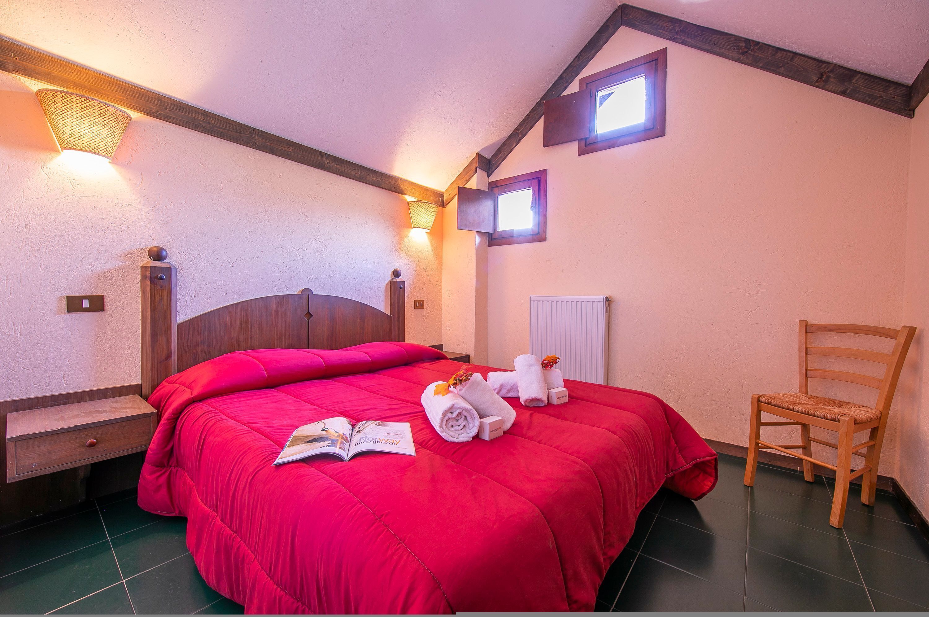 A bedroom with a large bed, wooden headboard, two wall-mounted lights, a wooden chair, and a small window.