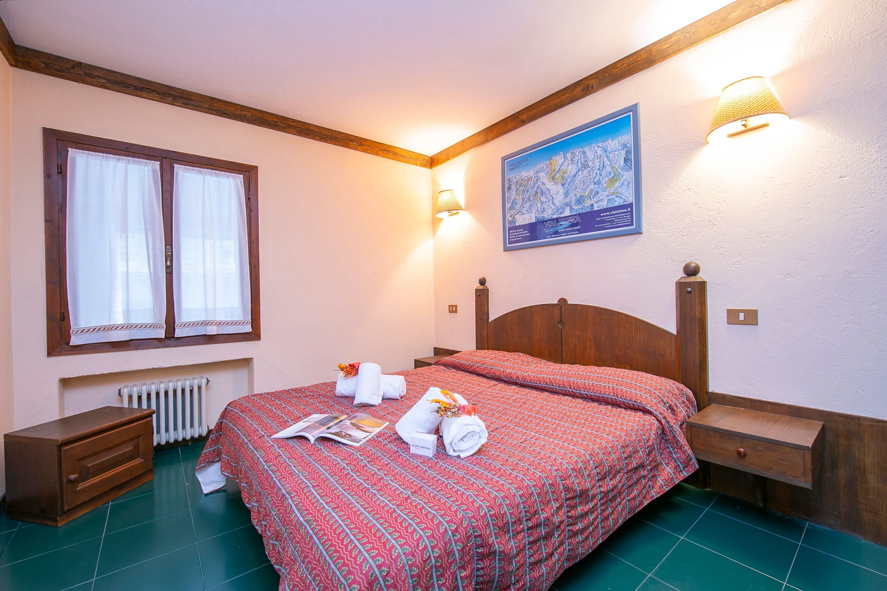 Una camera da letto con una testiera in legno, un letto con copriletto rosso fantasia, due comodini, un tavolino e una finestra con tende bianche. Si nota anche un termosifone e una mappa appesa al muro.